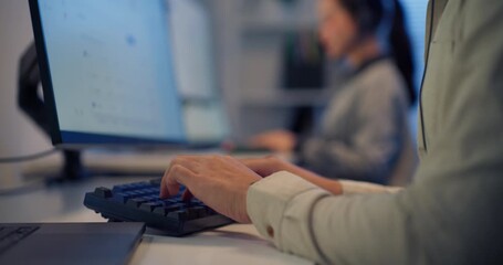 Close-up of Asian male call center agent typing on keyboard with headset in modern office at night, coworker in background — customer service, teamwork and communication. - Powered by Adobe