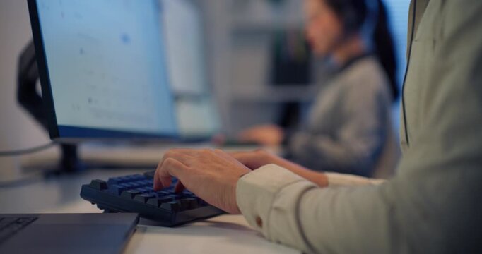 Close-up of Asian male call center agent typing on keyboard with headset in modern office at night, coworker in background — customer service, teamwork and communication.