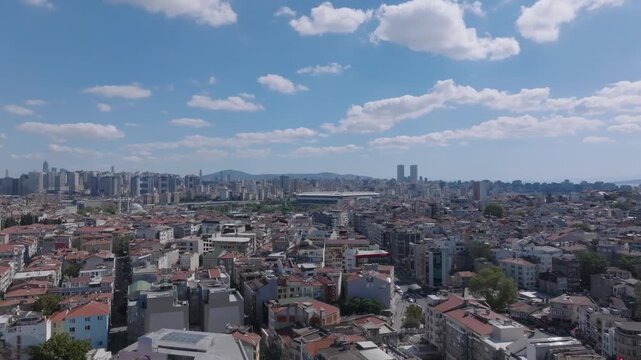 Aerial cityscape view of a coastal city, densely packed residential buildings with red terracotta rooftops, a large modern stadium on the left, tall skyscrapers in the distance, calm blue sea with yac