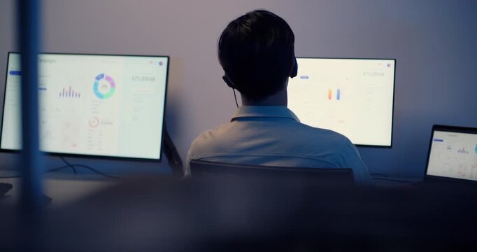 Rear view of Asian male call center service with headset working at night, using multiple computer screens for customer support, data analysis and communication in modern office.