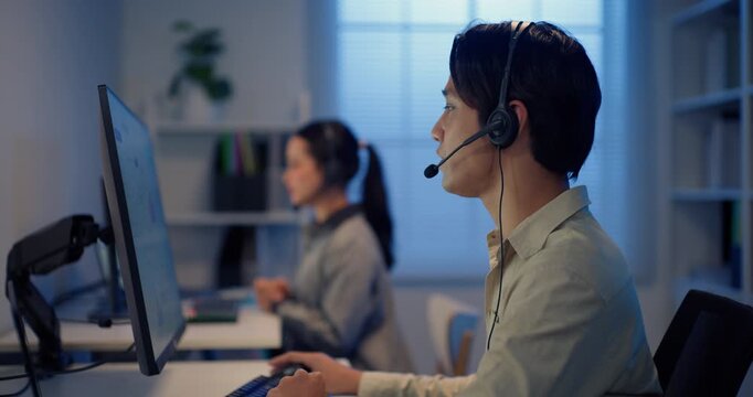 Asian male call center agent with headset working at night in a modern office, using computer for customer communication, IT support, business service and problem solving.