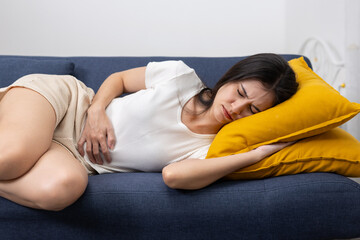 Asian women suffering from stomach pain or menstrual cramps while lying on a sofa, expressing...