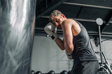 The white-skin man in a black sports uniform and white boxing gloves is boxing with a punching bag...