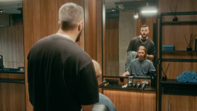 Medium rear shot of young Caucasian barber standing behind male customer sitting in chair, looking in mirror, discussing haircut request and style, then putting on protective cape