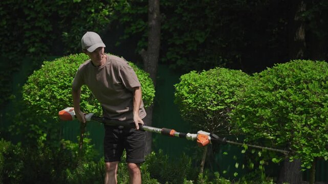 A man trims tall green shrubs with a long electric hedge trimmer in a well-maintained backyard. Dressed casually and wearing a cap, he focuses on shaping the foliage