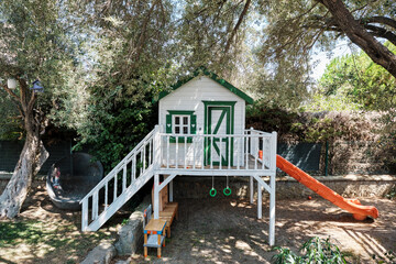 White and green playhouse with slide in a garden under olive trees