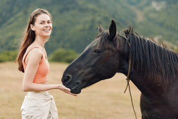 A smiling young woman in a peach top stands in an open field as a calm horse nuzzles her hand, capturing a serene moment of connection between human and animal.