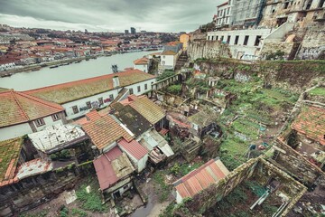 A cityscape view features dilapidated buildings and terraces overgrown with vegetation. The Douro River flows in the background, contrasting the aging structures with the lively city of Porto