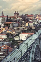 People walk across the upper level of the Dom Luis I Bridge in Porto. Old buildings with red tile roofs rise in the background on a cloudy day in Portugal