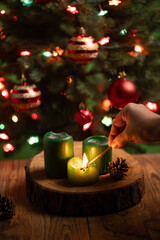 A hand lights a green candle using a matchstick on a wooden surface, next to unlit candles and pinecones. A Christmas tree and flashing lights are in the background.