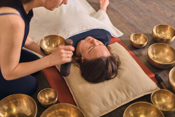 Young woman using singing bowls in serene space, calming lying adult with gentle strokes. Warm tone...
