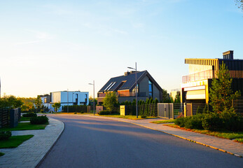 Modern houses along quiet street in evening light with greenery lining road on Curving street. Serene street features modern barn house, villa surrounded by lush greenery in warm light of evening.