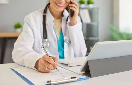 Doctor, nurse, medical professional at desk in office writing on clipboard, talking mobile phone, in white lab coat, stethoscope, focused work and multitasking, managing tasks and communication, call