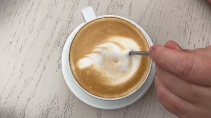 A cup of latte with latte art in the shape of a leaf or feather in the milk foam. A close-up of a beautiful coffee design. A spoon stirs coffee with milk foam. - Powered by Adobe