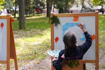 Child focused on painting at an easel adorned with autumn leaves in a park. The artwork process is underway, showcasing creativity and concentration amidst nature's colorful backdrop.