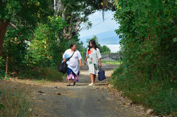 Fototapeta premium A relaxed walk through a village path as mother and daughter carry essentials, surrounded by tropical plants and mountain horizon in the distance.