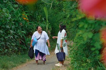 Fototapeta premium Two women stroll with laughter along a village path, carrying items while passing vibrant tropical flowers and enjoying their time outdoors.