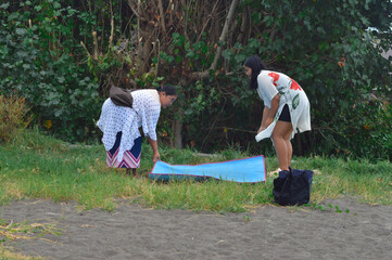 Obraz premium A mother and daughter carefully fold and pack their mat after finishing a picnic, surrounded by coastal grassland and sandy shore vegetation on a bright tropical day.