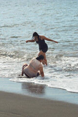 A teenage girl walks from shallow waves to shore while her mother rises from sitting at the waterâ€™s edge.
