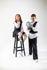 Obraz premium A young Caucasian girl sits on a stool, while a boy stands next to her, exploring a globe. Both wear formal attire, showcasing playfulness and curiosity.