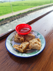 Tahu walik. Crispy browned tofu walik served in a natural wooden bowl with spicy sauce, with a blurry backdrop of a flowing river and rice fields. Menu in outdoor cafe.