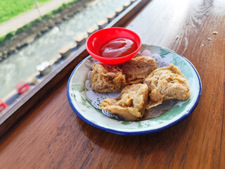 Tahu walik. Crispy browned tofu walik served in a natural wooden bowl with spicy sauce, with a blurry backdrop of a flowing river and rice fields. Menu in outdoor cafe.