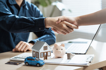 Handshake with house model, car, piggy bank, and coins on desk, symbolizing financial agreement, monthly payment, debt management, mortgage loan, and property investment.