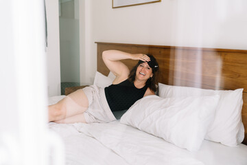 Obraz premium Smiling freelancer lifts sleep mask in bright room, symbolizing fresh start, work-from-bed comfort, and the ease of digital routines from home.