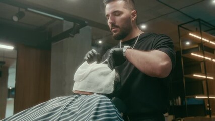 Medium shot of young Caucasian professional barber in black tshirt applying steamy hot white towel to face of mature Asian male patron, wrapping around and exposing nose, to open skin pores