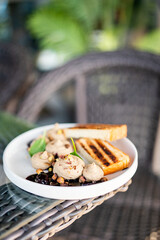 Gourmet appetizer with creamy spread, grilled bread, berry compote, and microgreens on white plate. Outdoor dining setup with wicker furniture and natural light.