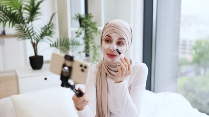 Young Muslim woman recording skincare vlog applying facial mask using brush in casual indoor setting. Promoting self-care, beauty, and skin health concepts with relaxed and motivating atmosphere.