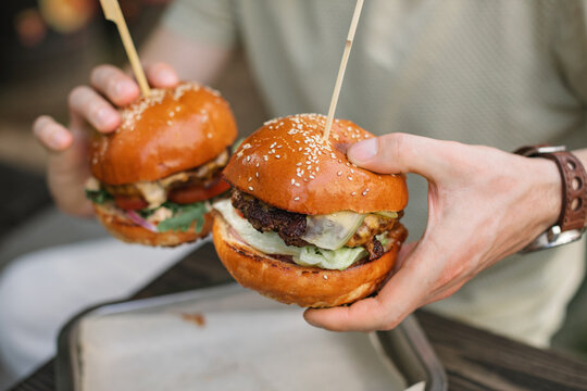 Close-up of a man sitting at a garden table holding two juicy cheeseburgers with pickles and salad
