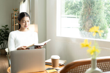 Young Asian woman enjoying a peaceful moment reading a book near window. Calm lifestyle, self-learning, and mindfulness at home.