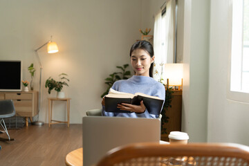 Young Asian woman enjoying a peaceful moment reading a book near window. Calm lifestyle, self-learning, and mindfulness at home.