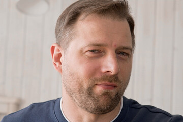 Fototapeta premium Handsome young man in blue jersey indoors. Male with stylish haircut, beard feel happy, calm and serious. Home interior background. Man looking to camera and into distance. Diversity People. Portrait