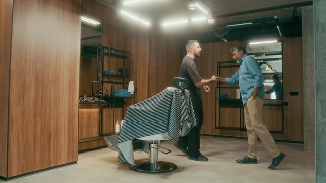 Wide shot of young Caucasian barber with tattoos standing in barbershop, cleaning tools, mature Asian male customer walking in, greeting with handshake, sitting in chair and discussing haircut request