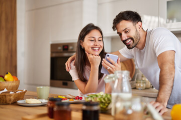 A couple smiles together in a bright kitchen, engaging with a smartphone as they share a lighthearted moment surrounded by tasty breakfast items like fruits and spreads.