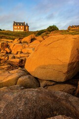 Granite rocks glowing in warm sunset light along the coast of Brittany, France. A historic seaside house with a turret stands in the background under a peaceful evening sky.