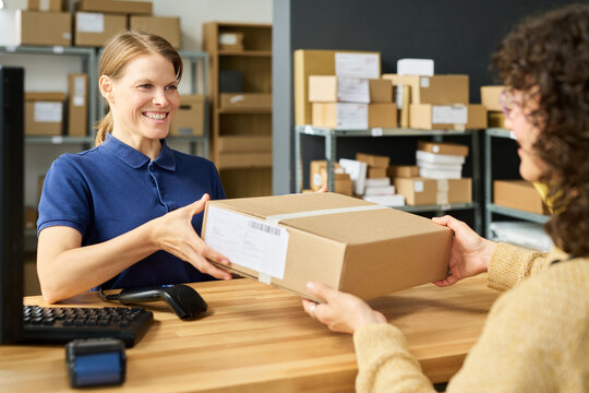 Caucasian young adult woman smiling while handing package to customer across counter in shipping or postal service office, shelves with parcels visible in background, barcode scanner on desk