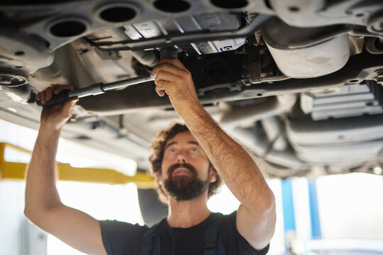A mechanic works under a car in a workshop, focused on tightening bolts with a wrench. The setting is bright and organized, showcasing various tools nearby.