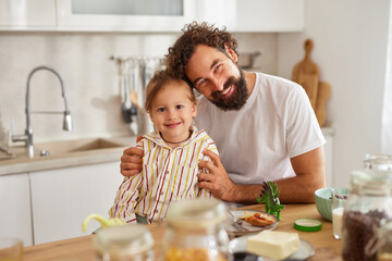A joyful father and his young child share a warm moment in the kitchen, smiling and enjoying their time together while preparing a meal. The scene is filled with light and warmth.