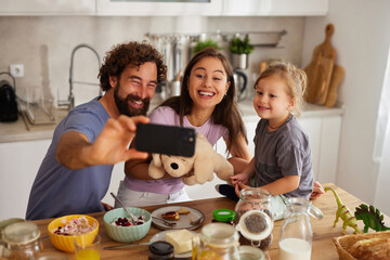 Smiling family sits at the kitchen table enjoying breakfast, taking a selfie with their pet stuffed animal, surrounded by food and love in a warm home setting.