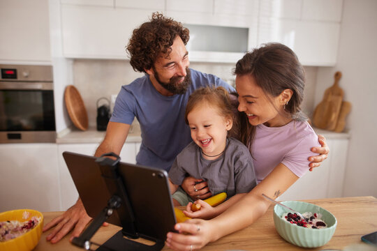 A happy family of three shares laughter and joy in their bright kitchen as they engage with a tablet. The parents embrace their young child, surrounded by food and warmth.