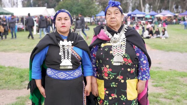 Two indigenous mapuche women in traditional clothing posing and talking at an outdoor cultural event