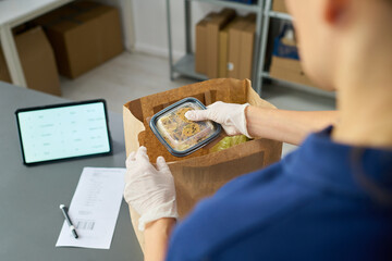 Young adult Caucasian woman wearing gloves packing food container into paper bag on table with digital tablet and printed receipt visible, preparing grocery delivery order in warehouse setting