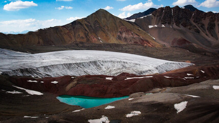 Aerial drone view of the grey ice tongue of the Lenin Glacier, red and brown mountains and a bright...
