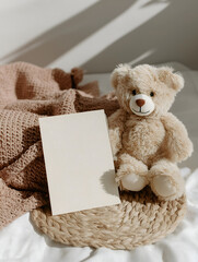 Cute teddy bear sitting beside a blank card on woven mat with soft blanket, perfect for baby shower invitations, nursery prints, greeting card mockups, and children’s product advertising.
