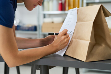 Young adult Caucasian woman writing order number on paper bag while checking printed list, leaning over table in workspace, focusing on organizing delivery or takeaway packaging process