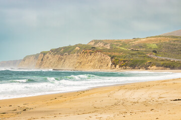 Wide sandy beach along San Mateo Coast State Beaches in California. Golden sand, coastal cliffs, ocean waves, and rolling hills create a peaceful natural seascape on the Pacific shoreline