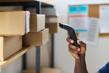 Black man hand holding barcode scanner scanning shipping label on cardboard box in warehouse storage area, focusing on logistics and inventory management process